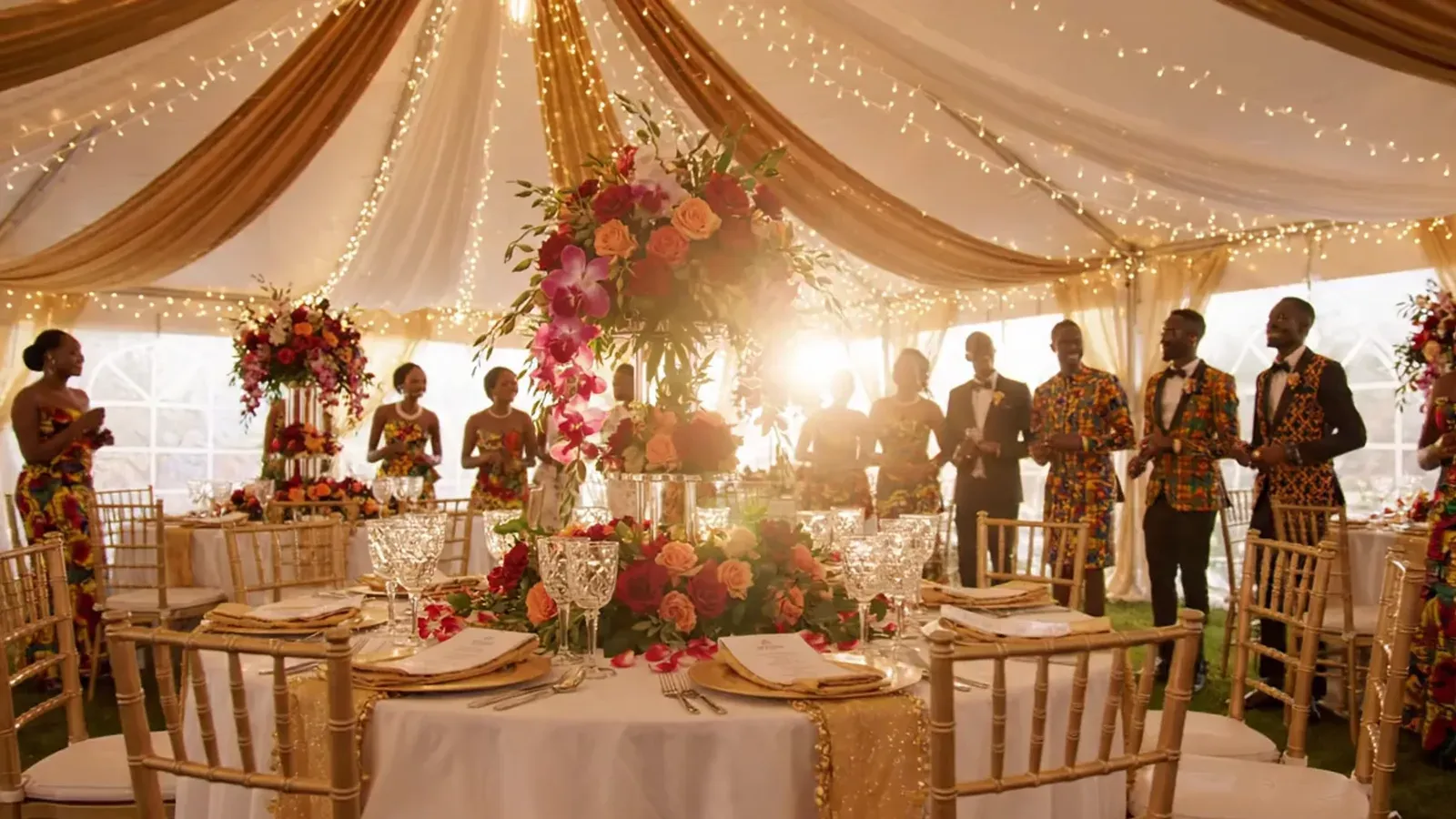 A marquee lit with fairy lights at golden hour, gold drapery overhead and long tables laid below.
