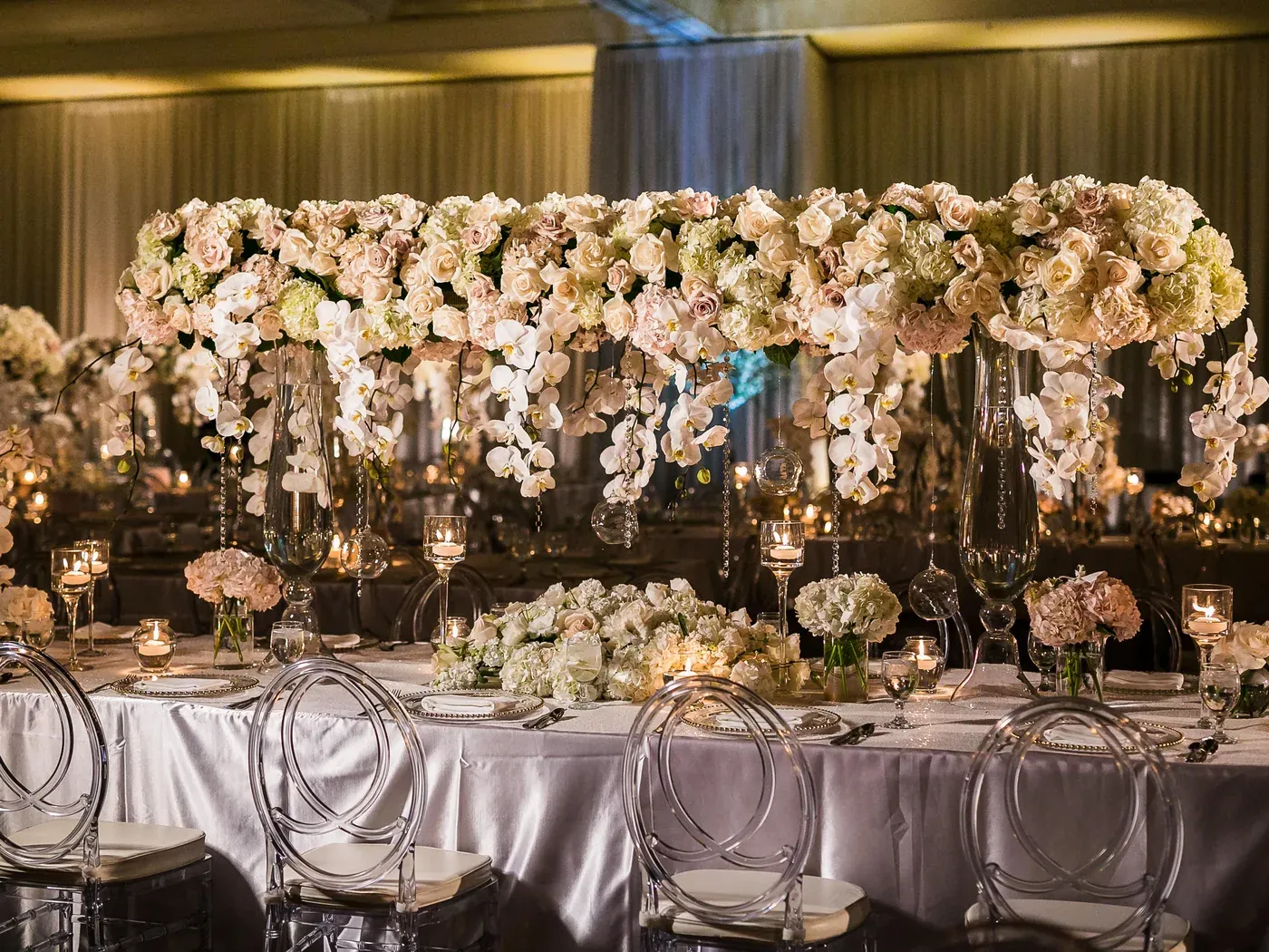A long raised floral installation of ivory roses and orchids above a top table set with ghost chairs and candles.